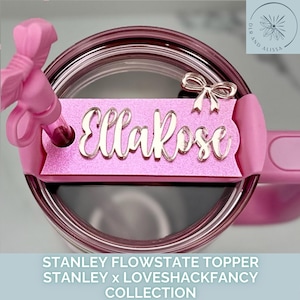 May include: A pink Stanley Flowstate topper with the name "EllaRose" in a cursive font. The topper has a glittery finish and a small bow accent. The image also shows a pink bow-shaped handle and the top of a clear tumbler.