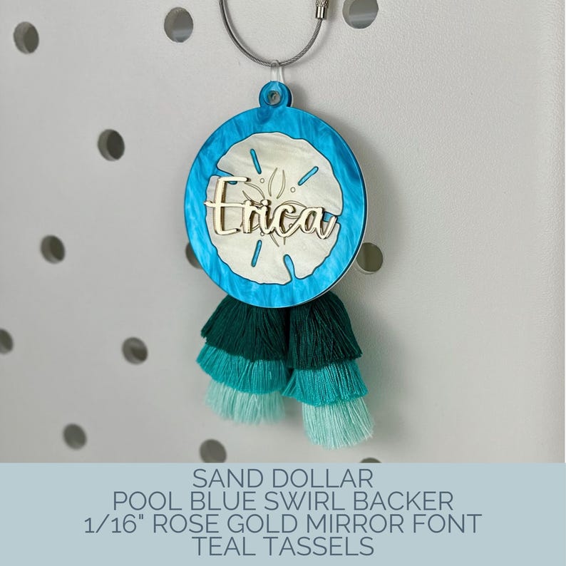 May include: A round, blue and white sand dollar-shaped keychain with the name "Erica" in rose gold. It has teal tassels and is attached to a silver keyring. The text on the image reads "SAND DOLLAR POOL BLUE SWIRL BACKER 1/16" ROSE GOLD MIRROR FONT TEAL TASSELS."