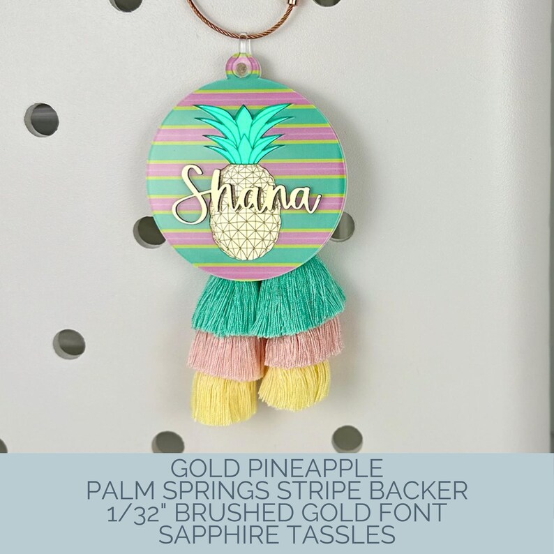 May include: A round, colorful keychain with a gold pineapple design and the name "Shana". The keychain has a striped background in shades of pink, green, and yellow, and is adorned with layered tassels in matching colors.