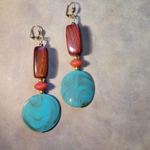 May include: A pair of dangle earrings featuring a combination of materials and colors. Each earring has a rectangular, reddish-brown wooden element, a small coral-colored bead, a gold-colored bead, and a large, round, turquoise-colored disc with a swirling pattern.
