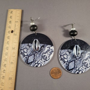May include: A pair of black and white snake print round earrings with a silver cowrie shell accent. The earrings are made of wood and have a black bead and a silver bead on the top. Shown next to a ruler, they are almost 4 inches long.