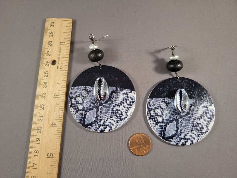 May include: A pair of black and white snake print round earrings with a silver cowrie shell accent. The earrings are made of wood and have a black bead and a silver bead on the top. Shown next to a ruler, they are almost 4 inches long.