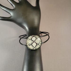 May include: A black metal bangle bracelet with a round, white and black snakeskin patterned glass cabochon.