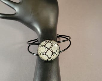 May include: A black metal bangle bracelet with a round, white and black snakeskin patterned glass cabochon.