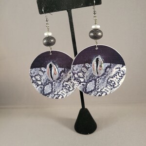 May include: A pair of large, round earrings with a black and white snake skin pattern. The earrings feature a silver shell charm and black wooden beads.