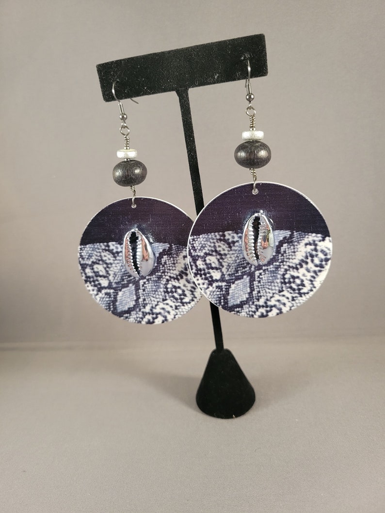 May include: A pair of large, round earrings with a black and white snake skin pattern. The earrings feature a silver shell charm and black wooden beads.