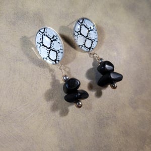 May include: Oval-shaped earrings with a white background and a black snake skin pattern. The earrings have a silver wire and a cluster of three black stones hanging from the bottom.