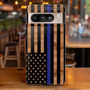 May include: A black phone case with a wood grain pattern and an American flag design with a blue line. The case has a camera lens cutout and a gold trim.