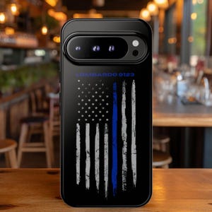 May include: A black smartphone case featuring a distressed American flag design in white with a single vertical blue stripe. The text LOMBARDO 0123 is printed in blue above the flag graphic. The case has a prominent horizontal camera bar.
