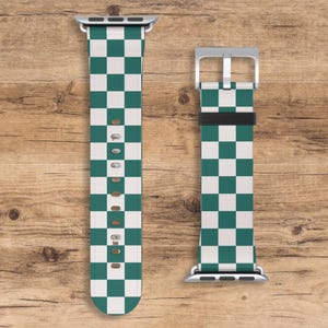 May include: Two watch bands with a green and white checkered pattern. The bands are made of a smooth material and have silver-colored clasps. The bands are displayed on a wooden surface.