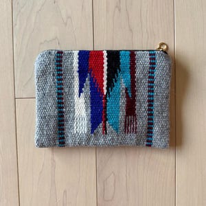 May include: A rectangular gray woven clutch with a colorful geometric design. The design features red, blue, black, and white patterns. The clutch has a gold-colored zipper and is made of a textured fabric.