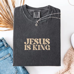 K&ouml;nnte beinhalten: Dunkelgraues Comfort Colors T-Shirt mit dem Text "JESUS IS KING" in beiger Schrift. Eine silberne Halskette mit Sonnenanh&auml;nger liegt auf dem Shirt. Das Shirt liegt auf einer wei&szlig;en Oberfl&auml;che mit einer Jeans und einem beigen Pullover.