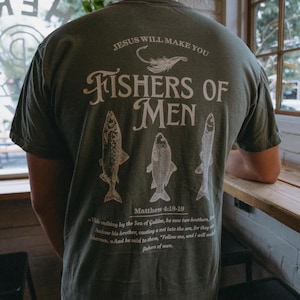May include: Olive green t-shirt with white text and illustrations. The back of the shirt features the phrase "FISHERS OF MEN" with fish illustrations and the Bible verse Matthew 4:18-19. The shirt also has the text "JESUS WILL MAKE YOU".