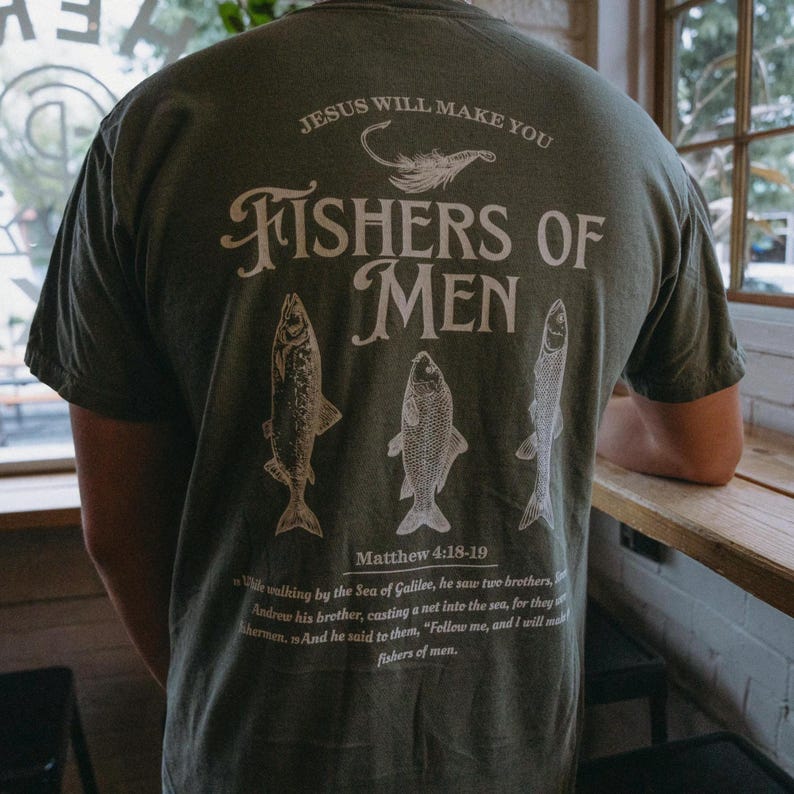 May include: Olive green t-shirt with white text and illustrations. The back of the shirt features the phrase "FISHERS OF MEN" with fish illustrations and the Bible verse Matthew 4:18-19. The shirt also has the text "JESUS WILL MAKE YOU".