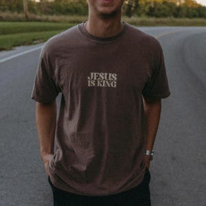 K&ouml;nnte beinhalten: Ein braunes T-Shirt mit dem Text "JESUS IS KING" in wei&szlig;er, stilisierter Schrift. Das Shirt hat kurze &Auml;rmel und einen Rundhalsausschnitt. Die Person tr&auml;gt eine Uhr am linken Handgelenk.