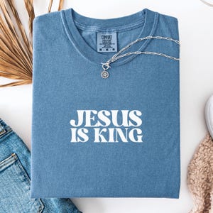 K&ouml;nnte beinhalten: Ein blaues T-Shirt mit dem wei&szlig;en Text "JESUS IS KING". Eine silberne Halskette mit Anh&auml;nger liegt auf dem T-Shirt. Das T-Shirt ist auf einer wei&szlig;en Oberfl&auml;che gefaltet, mit Jeans und einem beigen Pullover. Hergestellt von Comfort Colors.
