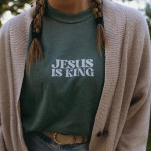 K&ouml;nnte beinhalten: Ein gr&uuml;nes Sweatshirt mit dem wei&szlig;en Text "JESUS IS KING". Das Sweatshirt wird mit einer beigen Strickjacke und einem braunen G&uuml;rtel getragen. Z&ouml;pfe sind auf beiden Seiten des Gesichts zu sehen. Der Stil ist l&auml;ssig.