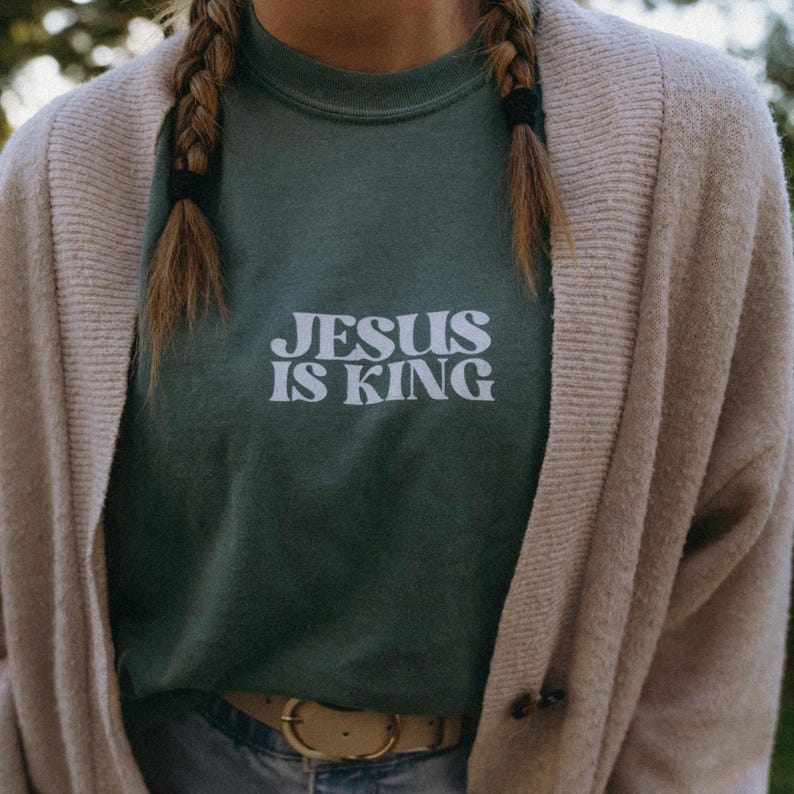 K&ouml;nnte beinhalten: Ein gr&uuml;nes Sweatshirt mit dem wei&szlig;en Text "JESUS IS KING". Das Sweatshirt wird mit einer beigen Strickjacke und einem braunen G&uuml;rtel getragen. Z&ouml;pfe sind auf beiden Seiten des Gesichts zu sehen. Der Stil ist l&auml;ssig.