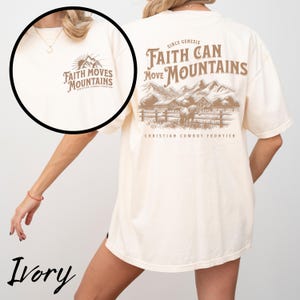 May include: Ivory-colored t-shirt with the text "Faith Moves Mountains" on the front and a mountain and horse graphic on the back. The back of the shirt also features the text "Faith Can Move Mountains" and "Christian Cowboy Frontier."