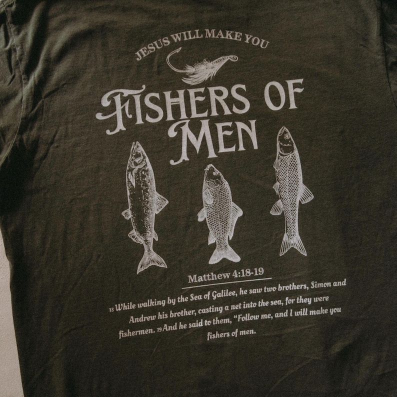 May include: Dark olive green t-shirt with a white graphic. The graphic includes the text "JESUS WILL MAKE YOU" above the words "FISHERS OF MEN" and three fish illustrations. Below the fish is the text "Matthew 4:18-19".