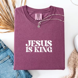 K&ouml;nnte beinhalten: Ein weinrotes Comfort Colors T-Shirt mit dem wei&szlig;en Text "JESUS IS KING". Eine silberne Halskette mit einem Sonnenblumenanh&auml;nger liegt auf dem Shirt. Das Shirt ist auf einer Jeans gefaltet.