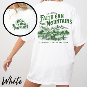 May include: White t-shirt with green text and graphics. The front features a small mountain graphic with the words "Faith Moves Mountains." The back displays a larger design with mountains, a fence, and horses, along with the text "Faith Can Move Mountains."