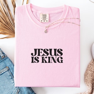 K&ouml;nnte beinhalten: Ein hellrosa T-Shirt mit dem Text "JESUS IS KING" in schwarzen Buchstaben. Eine silberne Halskette mit einem kleinen Anh&auml;nger liegt auf dem gefalteten Shirt. Das Shirt ist aus weichem Material.