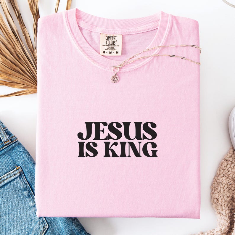 K&ouml;nnte beinhalten: Ein hellrosa T-Shirt mit dem Text "JESUS IS KING" in schwarzen Buchstaben. Eine silberne Halskette mit einem kleinen Anh&auml;nger liegt auf dem gefalteten Shirt. Das Shirt ist aus weichem Material.