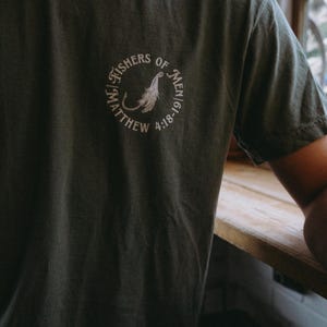 May include: Dark green t-shirt with a circular white graphic on the chest. The graphic includes the text "FISHERS OF MEN" and "MATTHEW 4:18-19" surrounding a stylized feather and fish hook design. The shirt is made of a soft material.