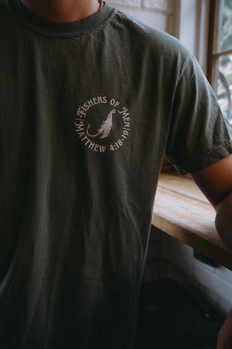 May include: Dark green t-shirt with a circular white graphic on the chest. The graphic includes the text "FISHERS OF MEN" and "MATTHEW 4:18-19" surrounding a stylized feather and fish hook design. The shirt is made of a soft material.