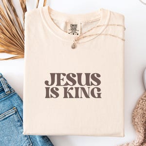 K&ouml;nnte beinhalten: Cremefarbenes T-Shirt mit dem Text "JESUS IS KING" in braunen Buchstaben. Das Shirt hat einen Rundhalsausschnitt und ist aus weichem Material. Eine goldene Halskette mit einem kleinen Anh&auml;nger liegt auf dem Shirt.