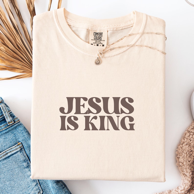 K&ouml;nnte beinhalten: Cremefarbenes T-Shirt mit dem Text "JESUS IS KING" in braunen Buchstaben. Das Shirt hat einen Rundhalsausschnitt und ist aus weichem Material. Eine goldene Halskette mit einem kleinen Anh&auml;nger liegt auf dem Shirt.