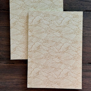 May include: Two cream-coloured paper cards with a gold wave pattern. A brown and tan feather and a small gold decorative object are on a dark wood surface.