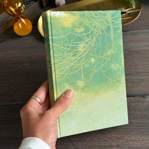 May include: A hardback book with a green and yellow gradient cover featuring a delicate, thin gold line illustration of tree branches. The book has a rectangular shape with a spine on the left side and a smooth, matte finish on the front cover.