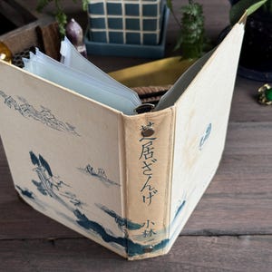 Könnte beinhalten: Ein cremefarbenes Fotoalbum mit einer Landschaftsillustration in blauer Tinte auf dem Einband und dem Buchrücken. Auf dem Buchrücken steht der japanische Text 芝居ざんげ und 小林. Das Album hat Metallecken, eine Ringbuchmechanik und innenliegende Klarsichthüllen für Fotos.