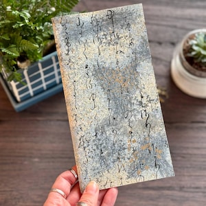 May include: A rectangular journal with a textured, gray and gold cover featuring black calligraphy. The journal is held in a hand, with plants in the background. The cover has a mottled appearance.