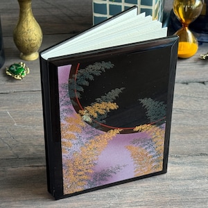 May include: A black hardcover book with a glossy finish featuring a decorative illustration of gold and green fern-like foliage against a lavender and black background with a thin red circular accent line and white paper pages visible at the top.