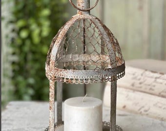May include: A distressed white metal lantern with a domed top and a decorative, openwork design. The lantern features a wire mesh and a round, white candle inside. The lantern is approximately 6 inches tall.