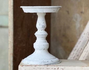 May include: A distressed white metal pedestal candle holder with a round top and a decorative base. The candle holder is placed on top of a stack of vintage books with handwritten text.