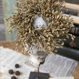 Puede incluir: Una corona floral seca de color marrón claro, con una pequeña base redonda blanca. La corona está sentada sobre un soporte negro, que está sobre una superficie blanca.