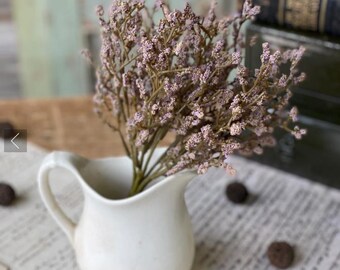 May include: A small, white ceramic pitcher holds a bouquet of dried, light purple flowers. The pitcher has a curved handle and a rounded body. The background includes vintage books and aged paper.