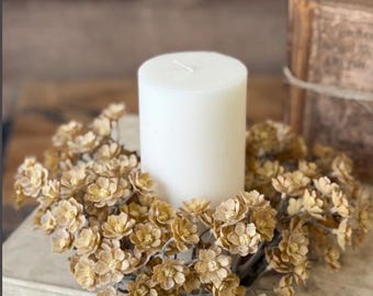 May include: A white pillar candle is centered on a stack of vintage books, surrounded by a wreath of small, dried, beige flowers. The candle and floral wreath create a rustic, natural aesthetic. The books are bound with twine.