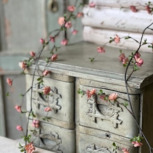 May include: A distressed, off-white cabinet with four drawers, decorated with pink floral garlands. The cabinet features ornate drawer pulls and is set against a soft green backdrop, creating a romantic, shabby-chic aesthetic.