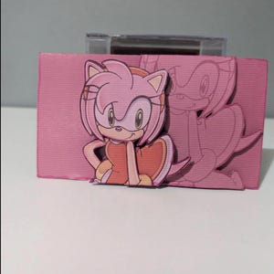 May include: A pink and white cardboard box with a cutout of a pink cartoon character with a red dress and pink hair. The character is holding a heart in her hand. The box has a clear plastic window on the top.