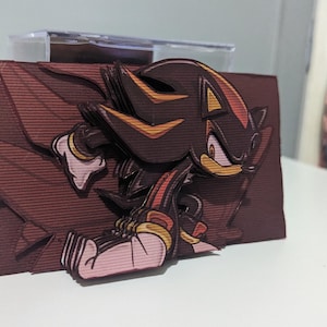May include: A papercraft figurine of Shadow the Hedgehog from the Sonic the Hedgehog video game series. The figurine is made of layered paper and features a 3D effect. Shadow is depicted in a running pose with his signature red and black attire.