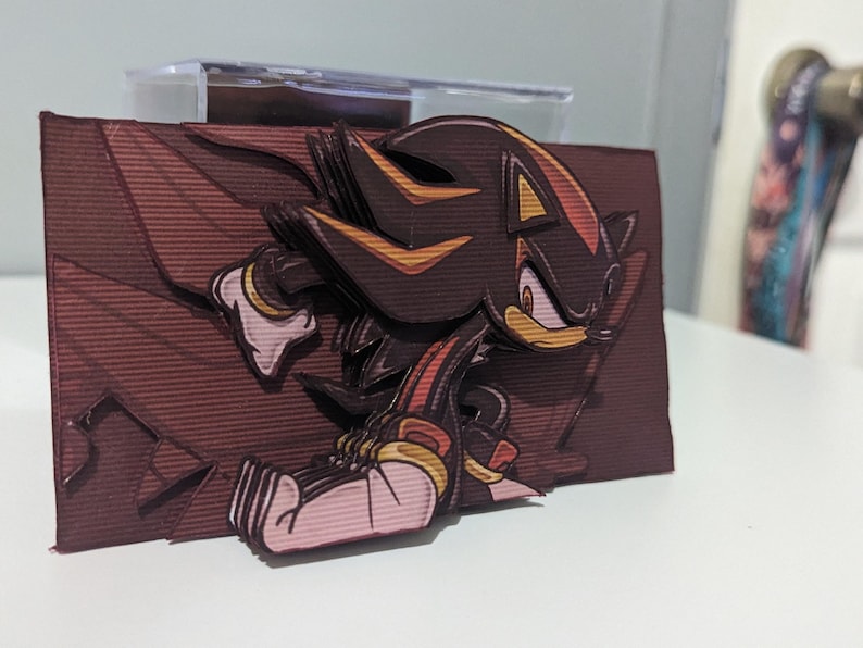 May include: A papercraft figurine of Shadow the Hedgehog from the Sonic the Hedgehog video game series. The figurine is made of layered paper and features a 3D effect. Shadow is depicted in a running pose with his signature red and black attire.