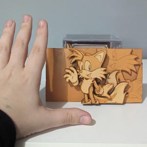 May include: A wooden cutout of Tails from the Sonic the Hedgehog video game series. The cutout is brown and features Tails's signature two tails. The cutout is displayed on a wooden base.