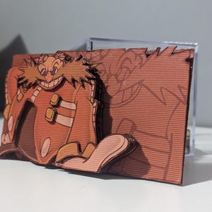 May include: A 3D papercraft model of Dr. Eggman, a villain from the Sonic the Hedgehog video game series. The model is made of brown cardboard and features a detailed depiction of Dr. Eggman's face and mustache.