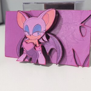 May include: A pink and purple papercraft figurine of a female character with bat wings. The character has a heart-shaped chest piece and a mischievous smile.