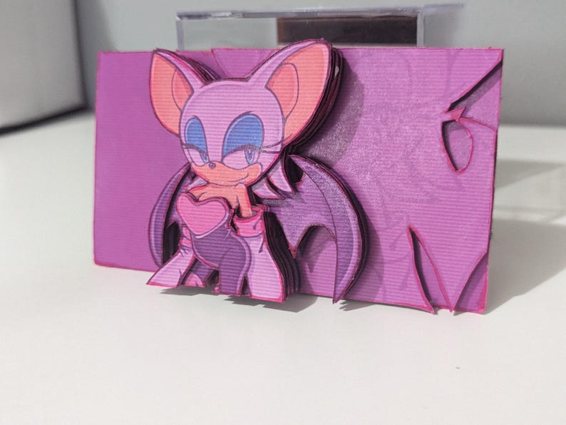 May include: A pink and purple papercraft figurine of a female character with bat wings. The character has a heart-shaped chest piece and a mischievous smile.